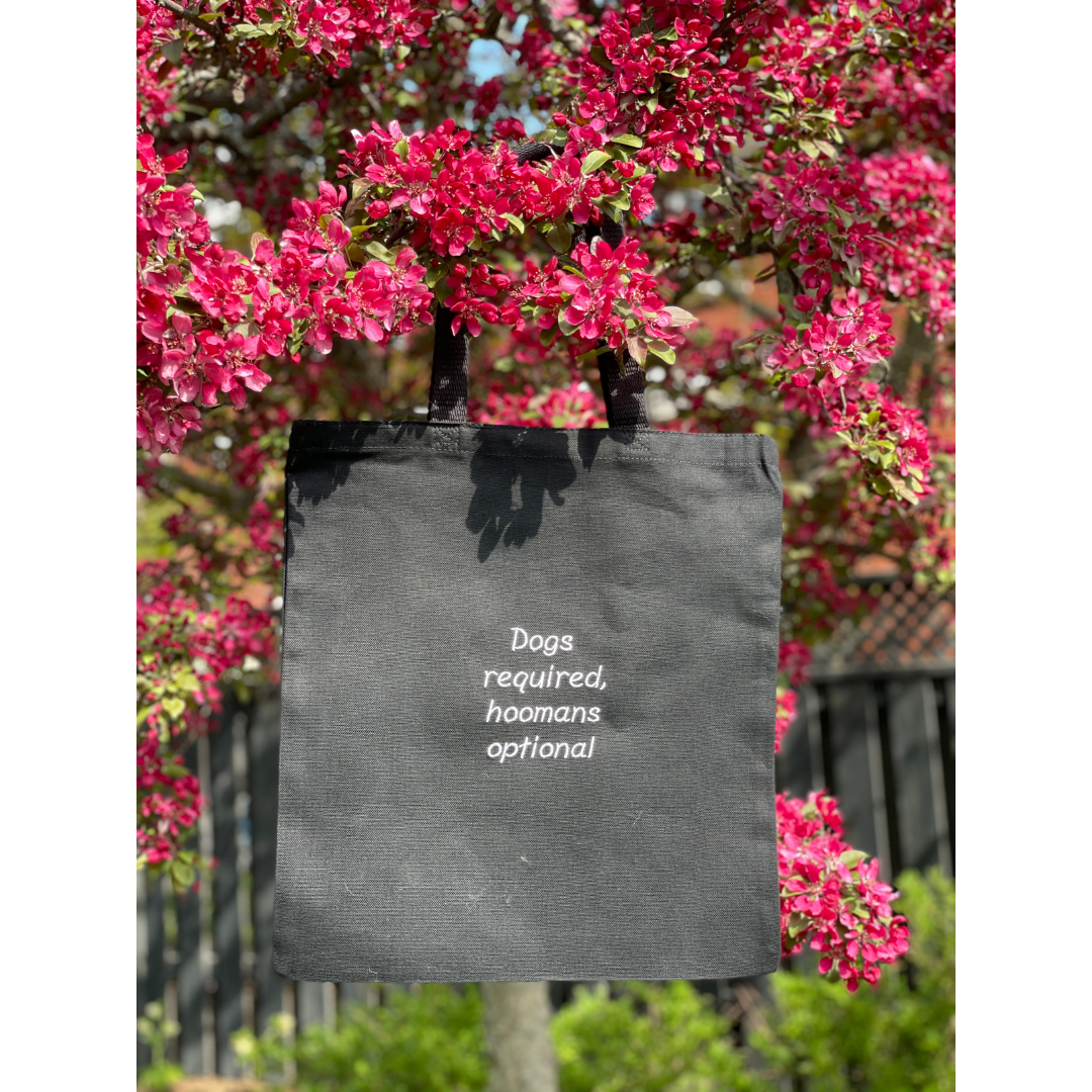 Black canvas tote bag hung on a tree. Quote on the bag says "Dogs required, hoomans optional"