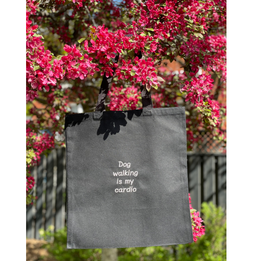 Black canvas tote bag hung on a tree. Quote on the bag says "Dog walking is my cardio"