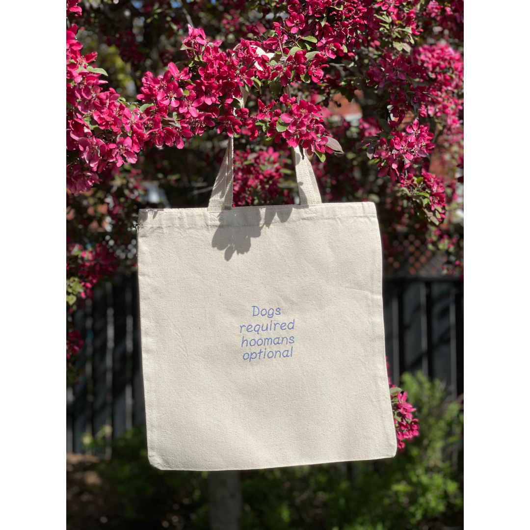 Natural canvas tote bag hung in a tree. Tote bag says "Dogs required, hoomans optional"