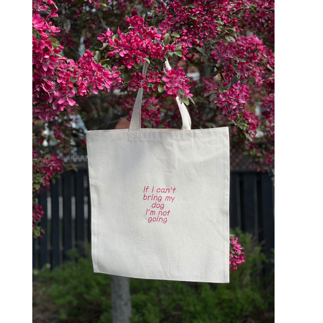 Natural canvas tote bag hung in a tree. Tote bag says "If I can't bring my dog I'm not going"