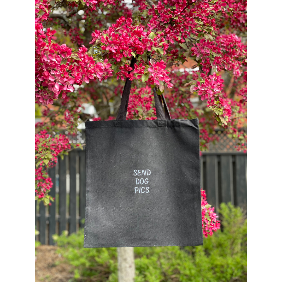 Black canvas tote bag hung on a tree. Quote on the bag says "SEND DOG PICS" all in capital letters
