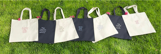 Black and Natural coloured canvas tote bags lying on the grass. Various quotes on the bags in different coloured fonts. font colours are white, black, light pink, fuchsia, and periwinkle. 